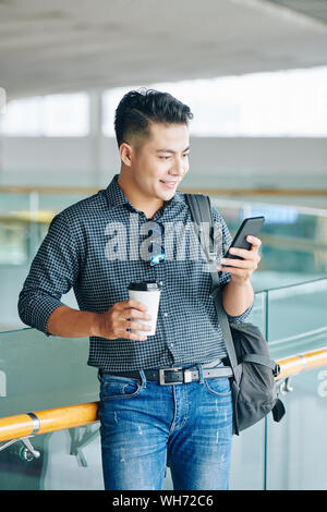 Smiling young Vietnamese man with backpack standing in airport terminal, drinking coffee and checking schedule in smartphone Stock Photo