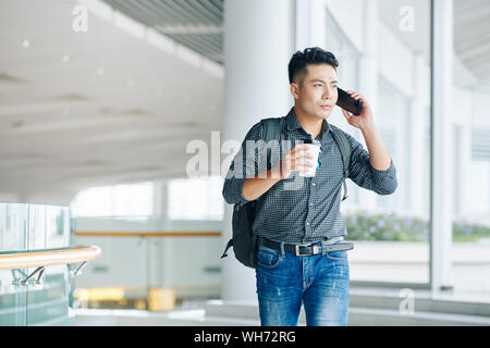 Handsome young Vietnamese man with backpack carrying cup of take away coffee and talking on phone when standing in office building Stock Photo