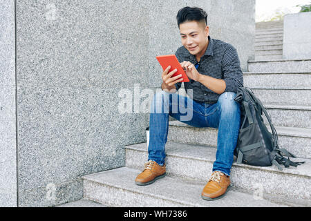 Smiling young Vietnamese student sitting on steps with backpack and using application on table computer Stock Photo