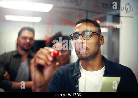 Business team looking at young businessman writing ideas and plans down on glass wall sticky notes and ideas Stock Photo