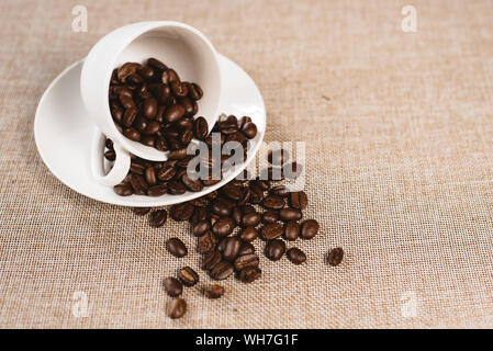 coffee beans overflow on a white cup over a burlap sack. concept of favorite drink and agriculture Stock Photo