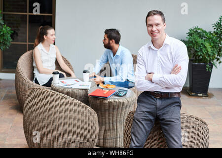 Portrait of confident young businessman folding arms and leaning on wicker cafe chair when his colleagues discussing project details in background Stock Photo
