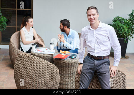 Handsome smiling entrepreneur in white short standing at cafe table and looking at camera when his colleagues having serious conversation in backgroun Stock Photo