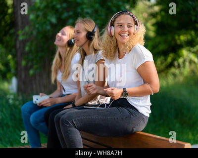 Three young women listening music with headphones and smartphones on a backrest of a bench Stock Photo