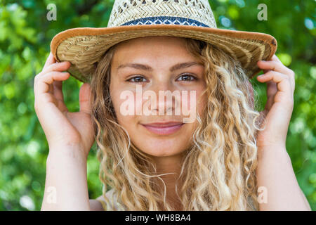 Portrait of smiling blond woman wearing straw hat, hands on hat Stock Photo