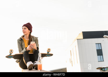 Stylish young woman with skateboard and cell phone sitting on a wall Stock Photo