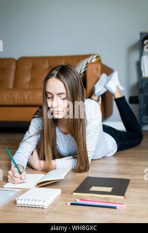 Young woman lying on the floor at home taking notes Stock Photo