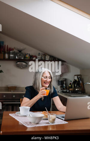 Portrait of laughing senior woman using laptop at breakfast table Stock Photo