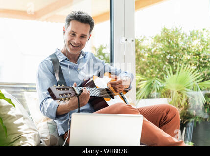 Mature man practising on the guitar, using laptop and earphones Stock Photo