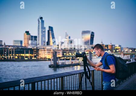 Young man photographing with tripod on embankment against urban skyline. Photographer holding smart phone with app. City life in London, United Kingdo Stock Photo