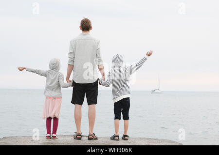 Back view of man standing in front of the sea with little daughter and son looking at distance Stock Photo