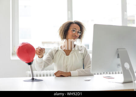 Portrait of young freelancer sitting at desk in office boxing punching ball while looking at computer Stock Photo