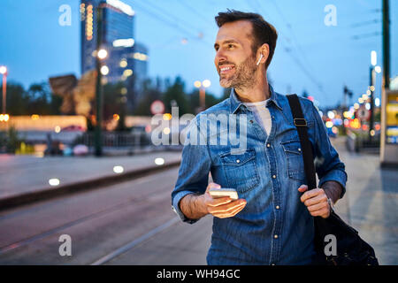 Smiling man using smartphone and listening to music through wireless headphones while waiting at tram stop during evening commute after work Stock Photo