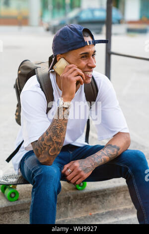 Portrait of tattooed young man on the phone sitting on his skateboard outdoors Stock Photo