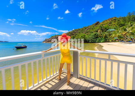 Woman in yellow dress on balcony of wooden jetty, looking at pristine white beach of Anse Gouvernement, near Cote d'Or Bay, in Praslin, Seychelles Stock Photo