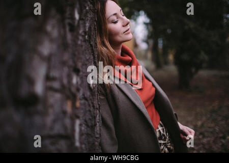 Portrait of woman with eyes closed leaning against tree trunk wearing turtleneck pullover Stock Photo