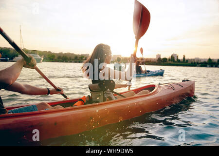 Happy young caucasian group of friends kayaking on river with sunset in the backgrounds. Having fun in leisure activity. Happy male and female model laughting on the kayak. Sport, relations concept. Stock Photo