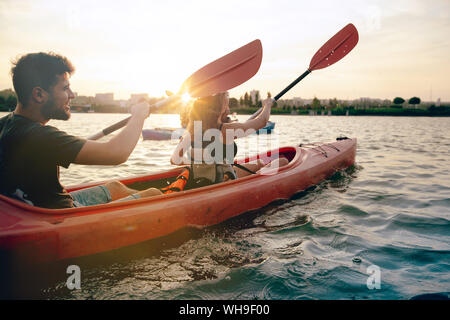 Confident young caucasian couple kayaking on river together with sunset in the backgrounds. Having fun in leisure activity. Romantic and happy woman and man on the kayak. Sport, relations concept. Stock Photo