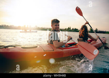Confident young caucasian couple kayaking on river together with sunset in the backgrounds. Having fun in leisure activity. Romantic and happy woman and man on the kayak. Sport, relations concept. Stock Photo