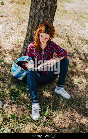 Redheaded motorcyclist leaning against tree trunk taking notes Stock Photo