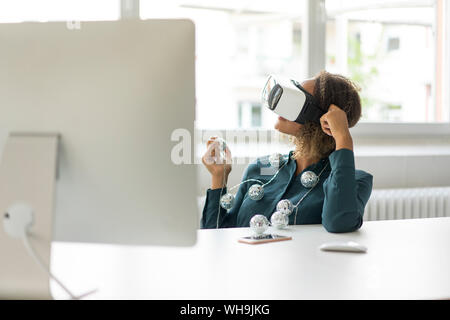 Young woman sitting at desk with chain of lights using Virtual Reality Glasses Stock Photo