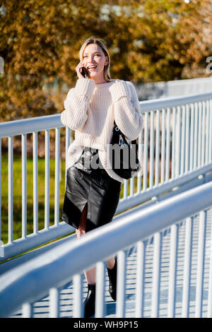 Smiling young woman on the phone walking on footbridge in autumn Stock Photo