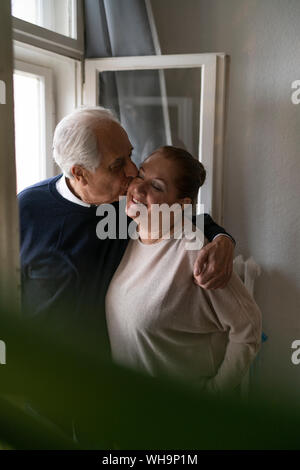 Happy senior couple kissing at the window at home Stock Photo