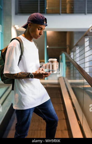Young man with backpack and coffee to go standing on escalator looking at cell phone Stock Photo
