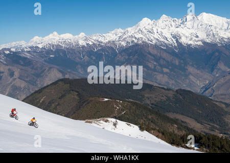 Mountain bikers cycle along a snow covered slope in the Himalayas with views of the Langtang range in the distance, Nepal, Asia Stock Photo