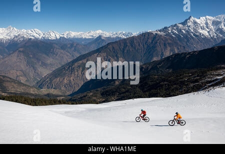 Mountain bikers descend a snow covered slope in the Himalayas with views of the Langtang range in the distance, Nepal, Asia Stock Photo