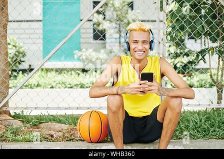 Portrait of happy laughing young Black basketball player sitting outdoors with smartphone in hands and enjoying music in headphones Stock Photo