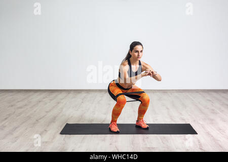 Portrait of young sporty healthy beautiful woman in black top and orange leggings doing squatting with elastic resistance band. Isolated, white wall, Stock Photo