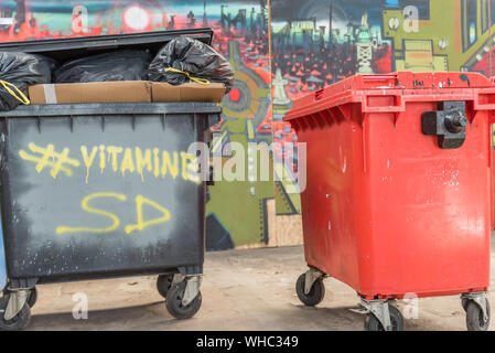 Black and red trash bins on wheels over flow links with fresh trash with graffiti against colorful background Stock Photo