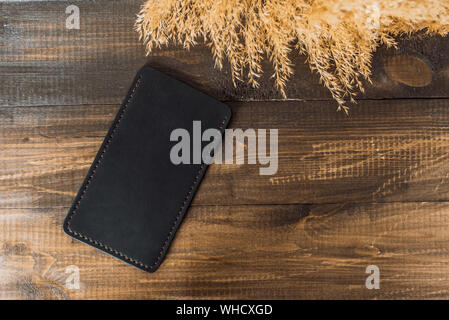 Expensive, beautiful cover of eco-leather on a background of wood and wheat. Beautiful nature care concept. Handmade phone case in black color. Stock Photo