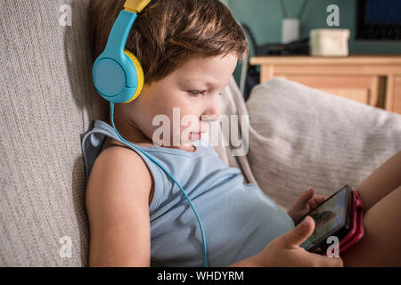 4 years boy in headphones watching videos with smartphone. Enjoying sitting on sofa in living room at home Stock Photo