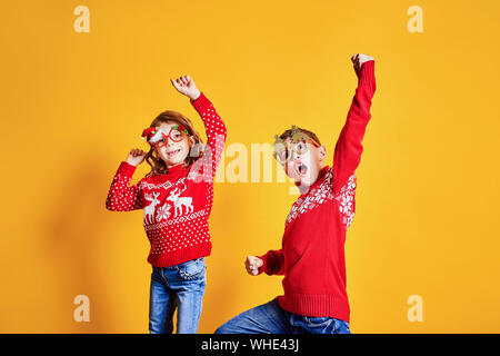 Confident children in warm red Christmas sweaters and decorated glasses looking at camera on yellow background Stock Photo