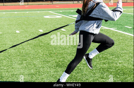 High school teenage girl is running fast while pulling a sled with weights across a green turf field. Stock Photo