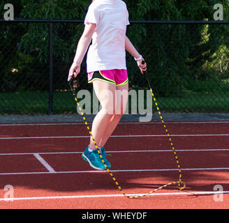 A teenage girl is using a black and yellow jump rope in the shade on a red track during practice on a warm day. Stock Photo