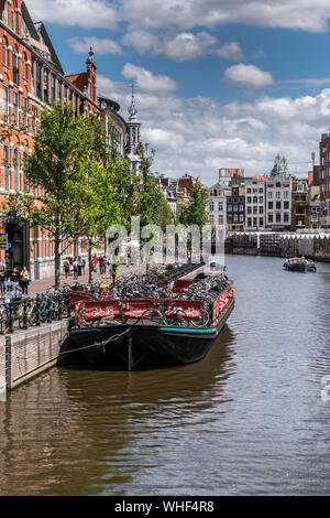 Amsterdam, the Netherlands - July 1, 2019: Dark flat barge is used as floating bike parking on Singel canal under blue-gray cloudscape. Munt tower spi Stock Photo