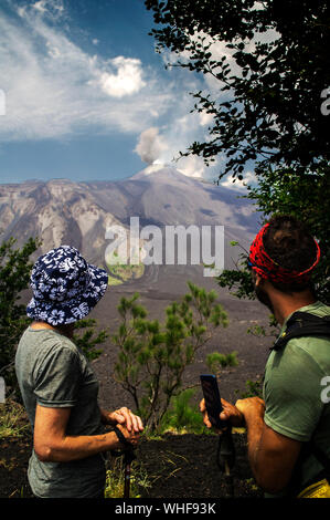 Two hikers watch the active crater of Mount Etna. Gases coming out seem to merge with the clouds.  Old and new lava flows in different shades of grey. Stock Photo
