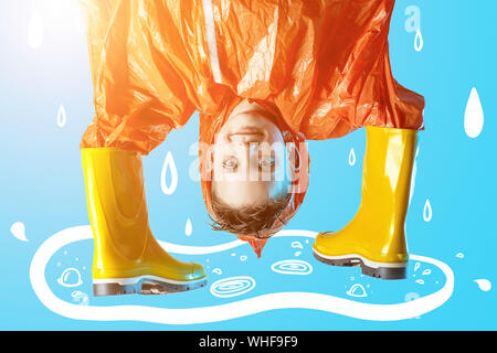 smiling boy in orange raincoat stuck his hands in rubber boots on a blue background Stock Photo