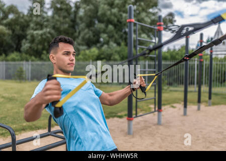 Athlete guy does pectoral muscle wiring, trains in summer city, holds loops his hands, sports straps for push-ups pull-ups, fitness, workout. Sportswe Stock Photo