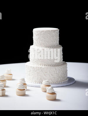 Three tiered wedding cake and cupcakes on table with black background Stock Photo