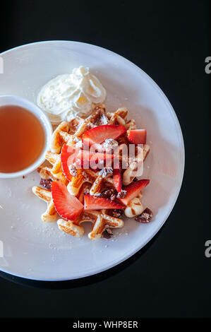 Crispy waffle with fresh strawberries baked walnuts whipping cream and honey in white plate on black table. Top view food image Stock Photo