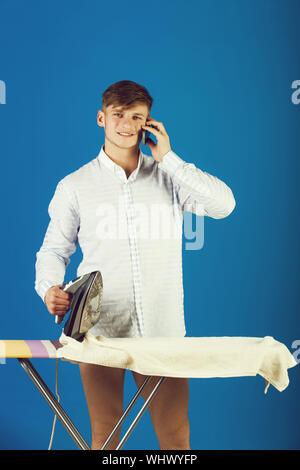 Macho wearing striped shirt. Man ironing laundry on board. Guy talking on mobile phone on blue background. Model using iron. Housework and technology Stock Photo