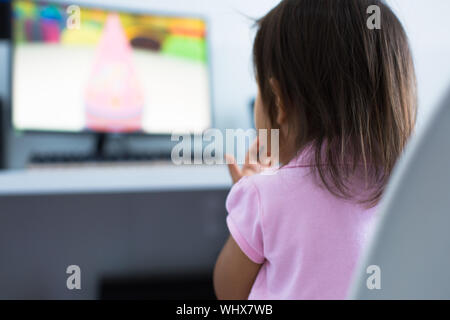 A 3-5 years old girl toddler sitting at the computer desk watching cartoon shows on the monitor at home. Children educational and development concept. Stock Photo