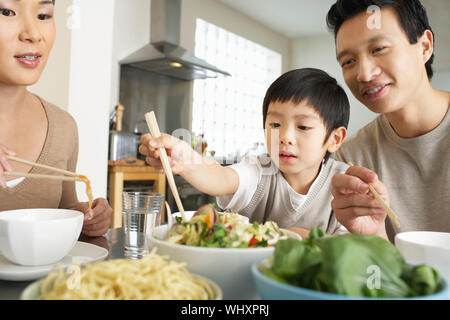 Young parents watching son trying to use chopsticks at dining table Stock Photo