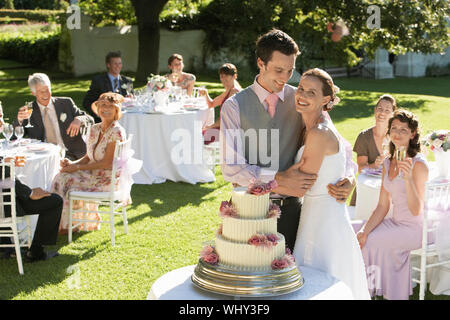 Happy young bride and groom in front of wedding cake while guests sitting in garden Stock Photo