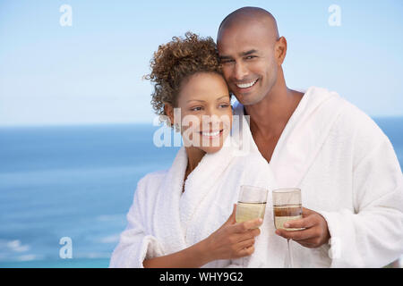 Portrait of happy young couple in bathrobes holding drinks with ocean in background Stock Photo
