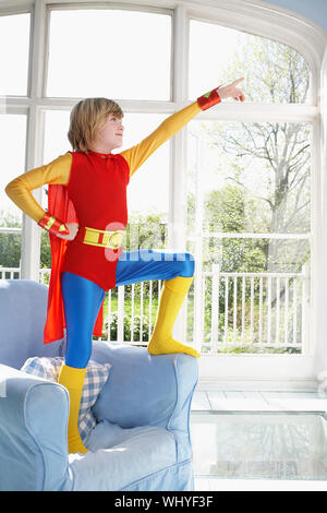 Full length of a young boy in superman costume standing on armchair and pointing up Stock Photo